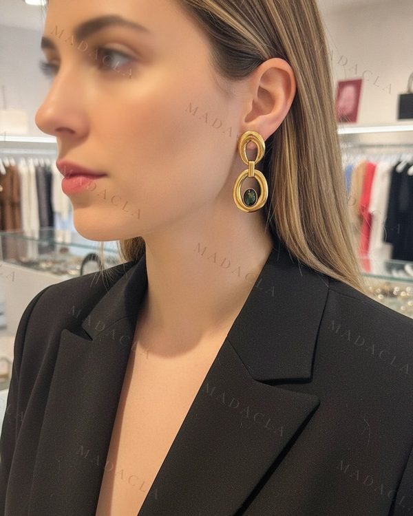 Pendientes dorados, largos, con una pieza ovalada superior. De ella cuelga otro aro ovalado con un cabujón verde oscuro.