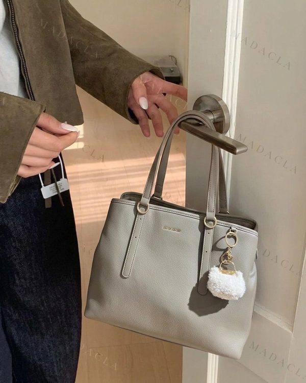 Bolso grande gris con dos asas de mano, detalles metálicos dorados, tiras decorativas verticales y un llavero colgante con pompón blanco. Se venden por separado.