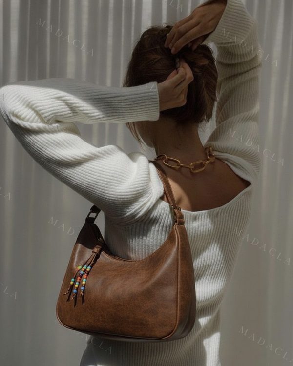 Bolso de hombro marrón de corte redondeado, con asa corta y un adorno colgante de cordones con cuentas de colores en un lateral.