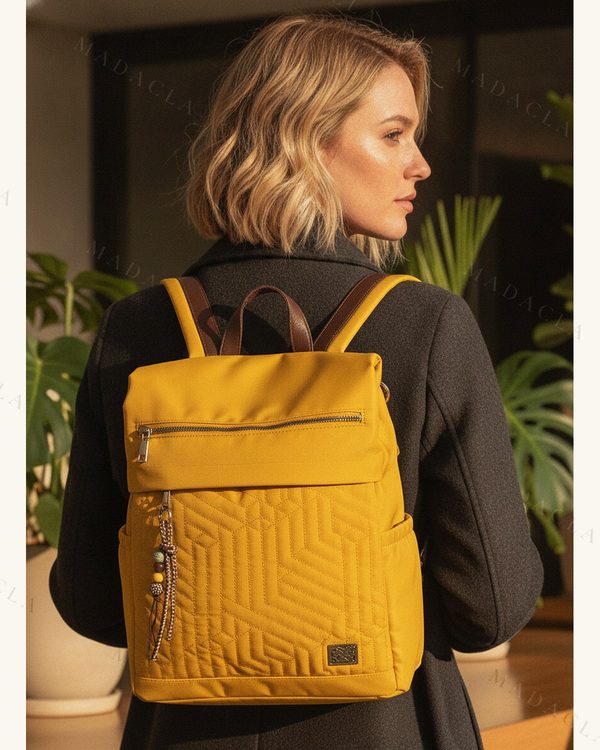 Mochila de color amarillo mostaza con bolsillos exteriores, cremalleras metálicas, asa de mano superior, correas marrones y un pequeño adorno colgante de cuentas.
