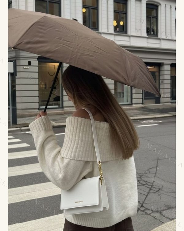 Bolso blanco rectangular con textura granulada y solapa. Tiene un asa fina con detalles de cadena dorada y letras doradas visibles.