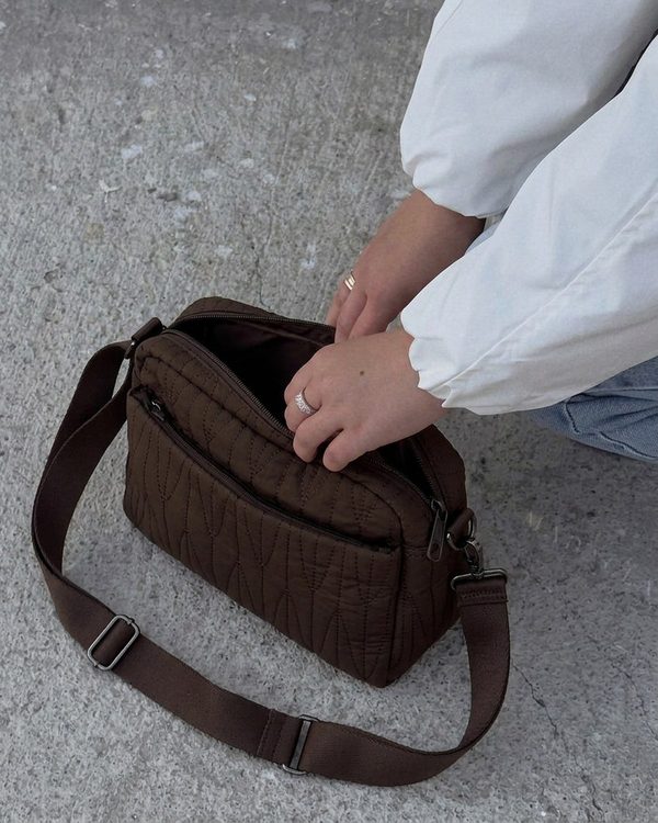 Bolso bandolera marrón oscuro de tejido acolchado con patrón de ondas y cremalleras frontales. Incluye correa ajustable con hebillas metálicas.