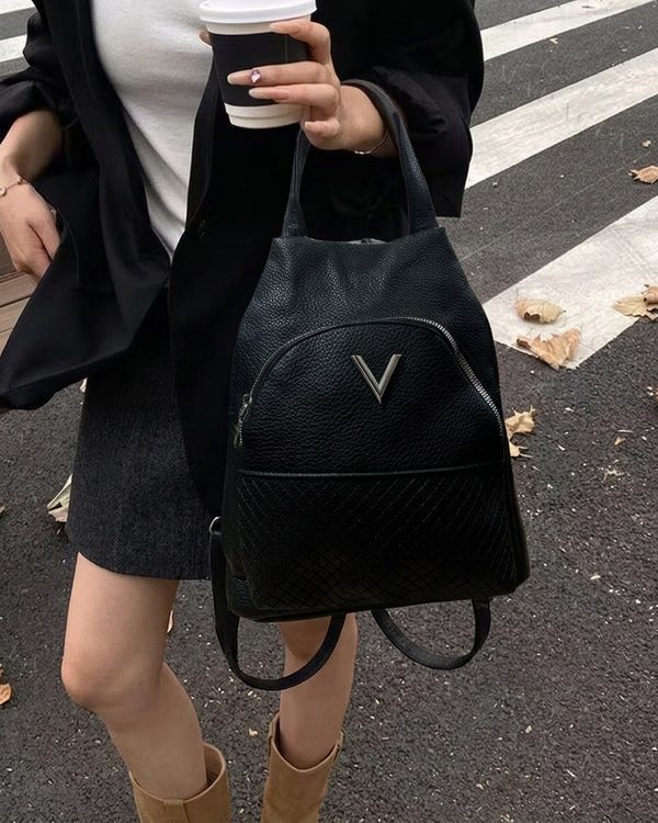Mochila negra de textura granulada con cierre de cremallera y bolsillo frontal. Incluye adorno metálico plateado y detalle de rombos en la parte inferior.
