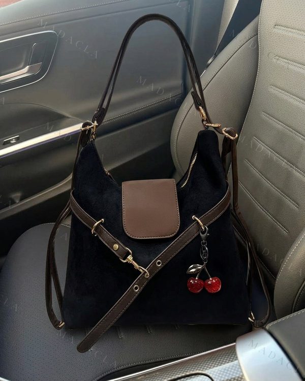 Bolso de hombro negro de tejido suave aparente con solapa y correas marrones, y herrajes dorados. Adorna un colgante de dos cerezas rojas brillantes. Se venden por separado.