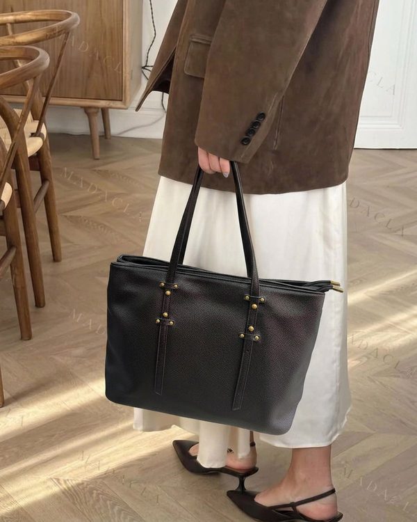 Bolso tote rectangular negro de textura granulada con asas dobles. Presenta remaches dorados y tiras verticales decorativas en el frontal, y cierre de cremallera superior.