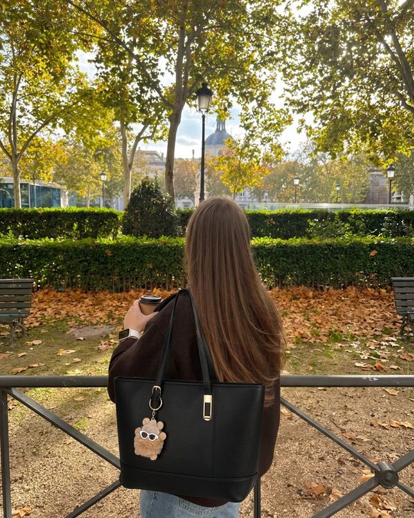 Bolso tote negro liso de asas de hombro. Incluye una pieza metálica dorada vertical en el frente y hebilla dorada decorativa.