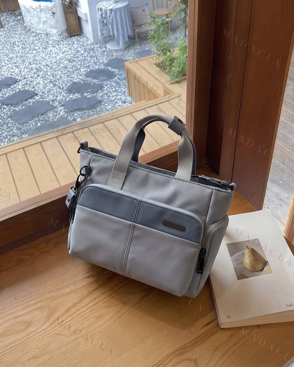 Tote pequeño de tejido en gris claro, con detalles en gris oscuro, asas cortas, bolsillo frontal y lateral con cremallera.
