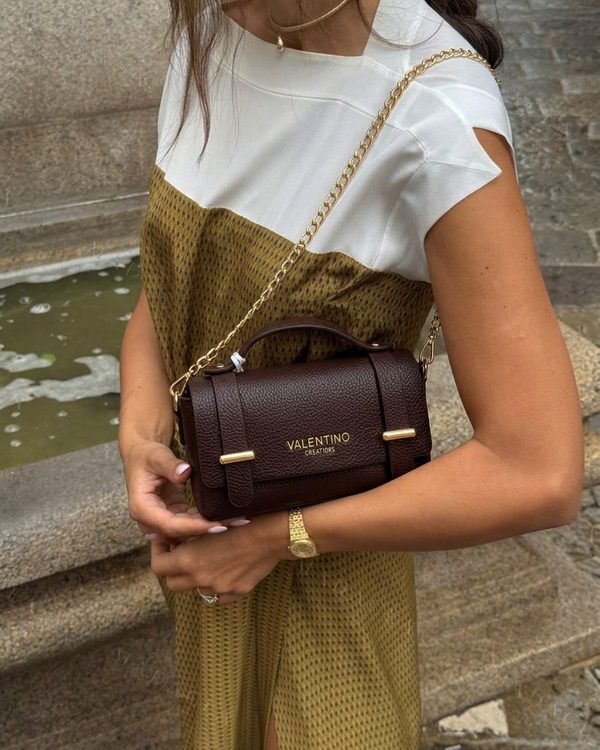 Bolso pequeño estructurado en marrón oscuro de aparente piel granulada, con asa superior, cadena dorada y cierre frontal con detalles metálicos, letras y logos.