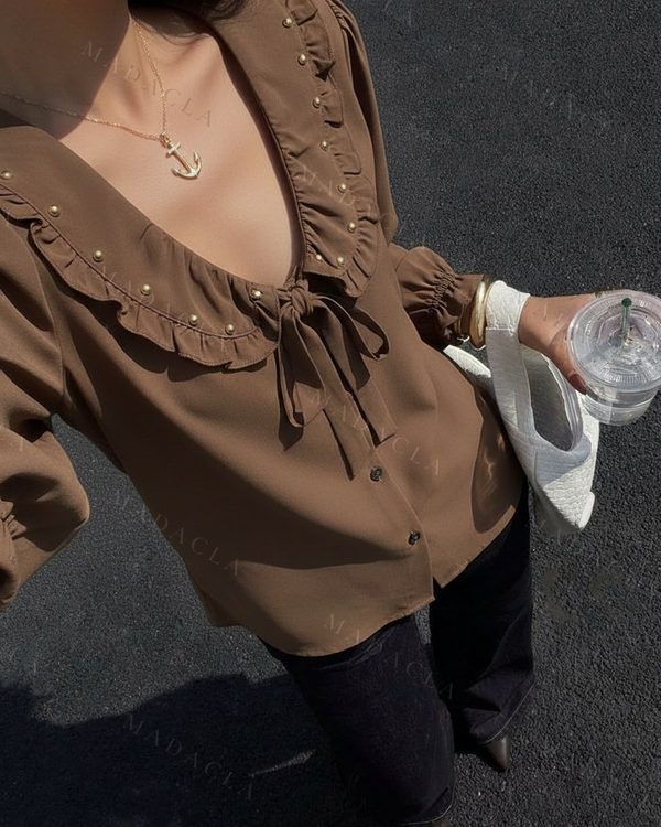 Blusa marrón de tejido fluido con escote pico, lazo y botones frontales. Su cuello de volantes está adornado con remaches dorados; mangas largas elásticas.