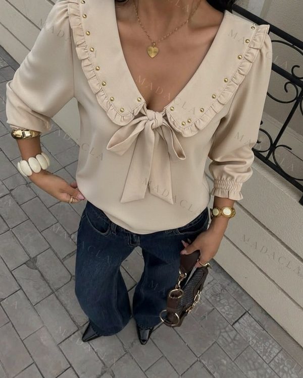 Blusa beige de tejido fluido, escote en V con lazada y cuello de solapas fruncidas con remaches dorados. Mangas largas con puños elásticos.