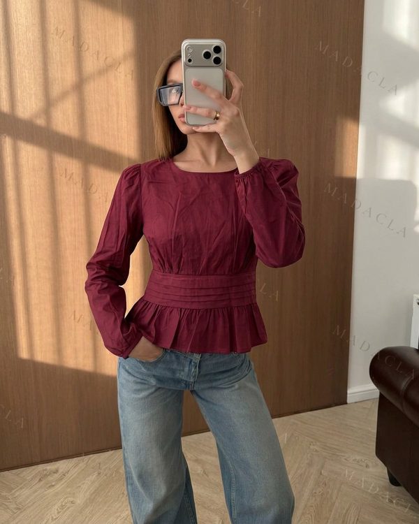 Camisa granate de corte peplum con escote redondo y mangas largas abullonadas. Presenta pliegues en el talle y fruncidos en el bajo.