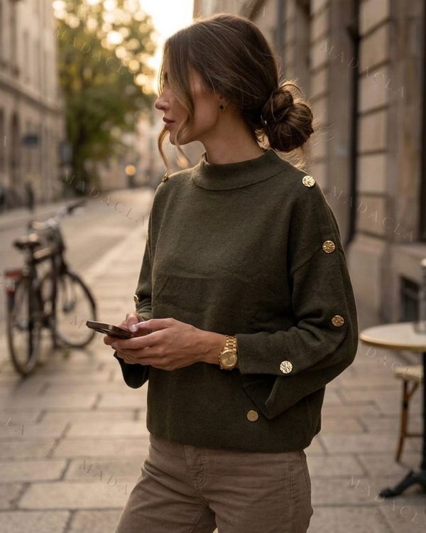 Jersey de punto fino verde oscuro, cuello alto y corte recto. Lleva botones dorados en hombros y puños acampanados con aberturas.
