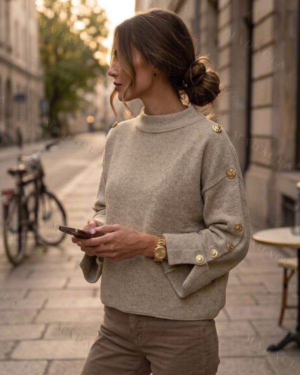Jersey de punto fino beige, cuello alto y corte recto. Lleva botones dorados en hombros y puños acampanados con aberturas.