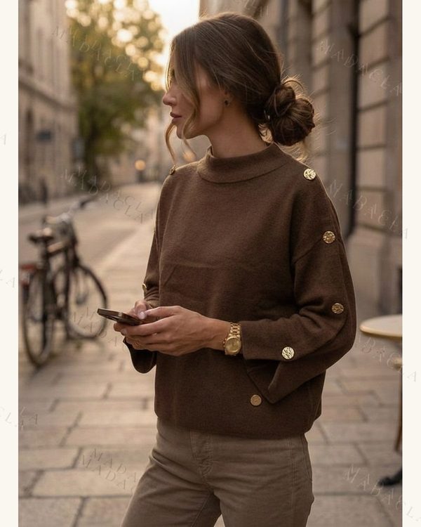 Jersey de punto fino marrón, cuello alto y corte recto. Lleva botones dorados en hombros y puños acampanados con aberturas.