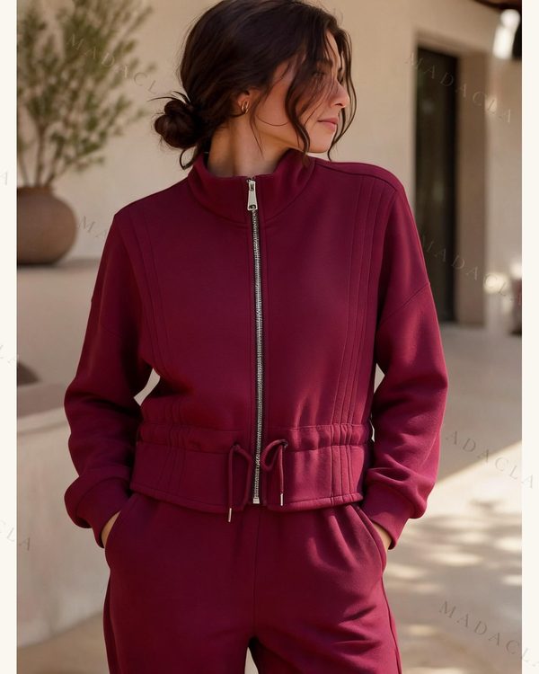 Chaqueta deportiva burdeos con cuello alto y cremallera plateada central. Lleva manga larga, puños elásticos y cordón ajustable en la cintura.