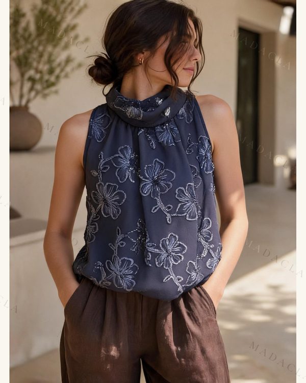 Blusa azul oscura sin mangas, con cuello alto vuelto y bordados de flores en tono grisáceo con brillo por toda la prenda.