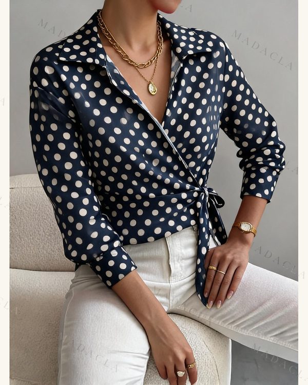 Blusa azul oscura de manga larga con lunares blancos, cuello con solapas, escote de pico cruzado y lazada lateral en la cintura.