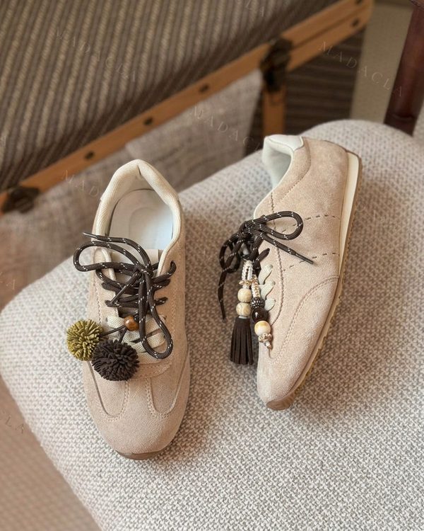 Deportivas beiges de cordones marrones estampados. Llevan borlas, abalorios y pompones colgando en los cordones. Suela plana y puntera redondeada.