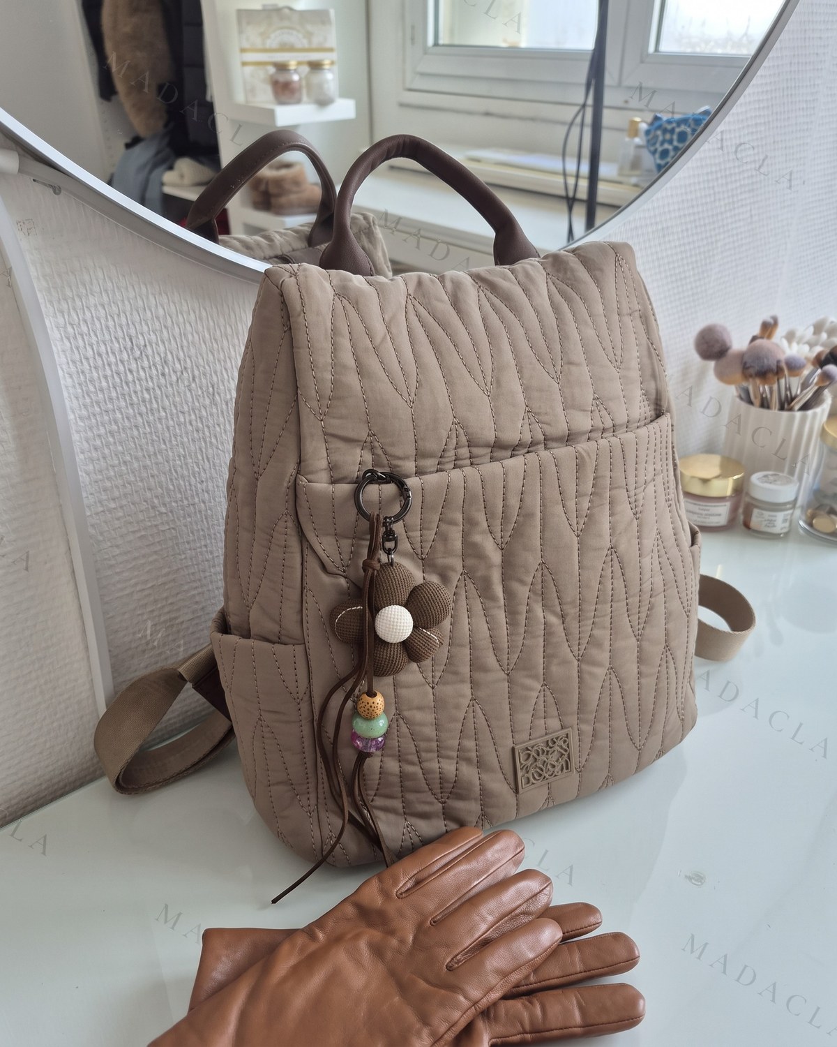 Mochila acolchada con pespuntes en zigzag. Asa de piel marrón arriba, tirantes anchos y bolsillos laterales. Lleva colgante de flor de tela con cuentas de colores.