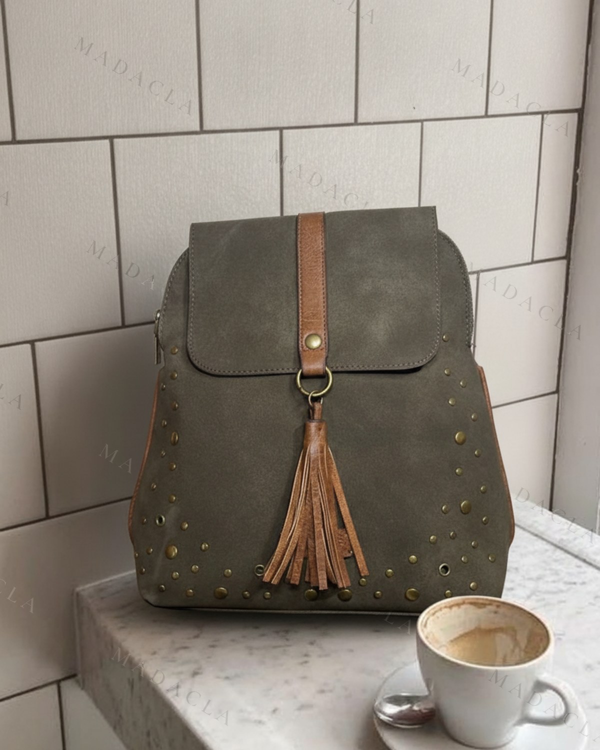 Mochila en verde caqui efecto ante, con tachuelas de bronce por todo el cuerpo, solapa de cierre y borla de piel marrón colgando.