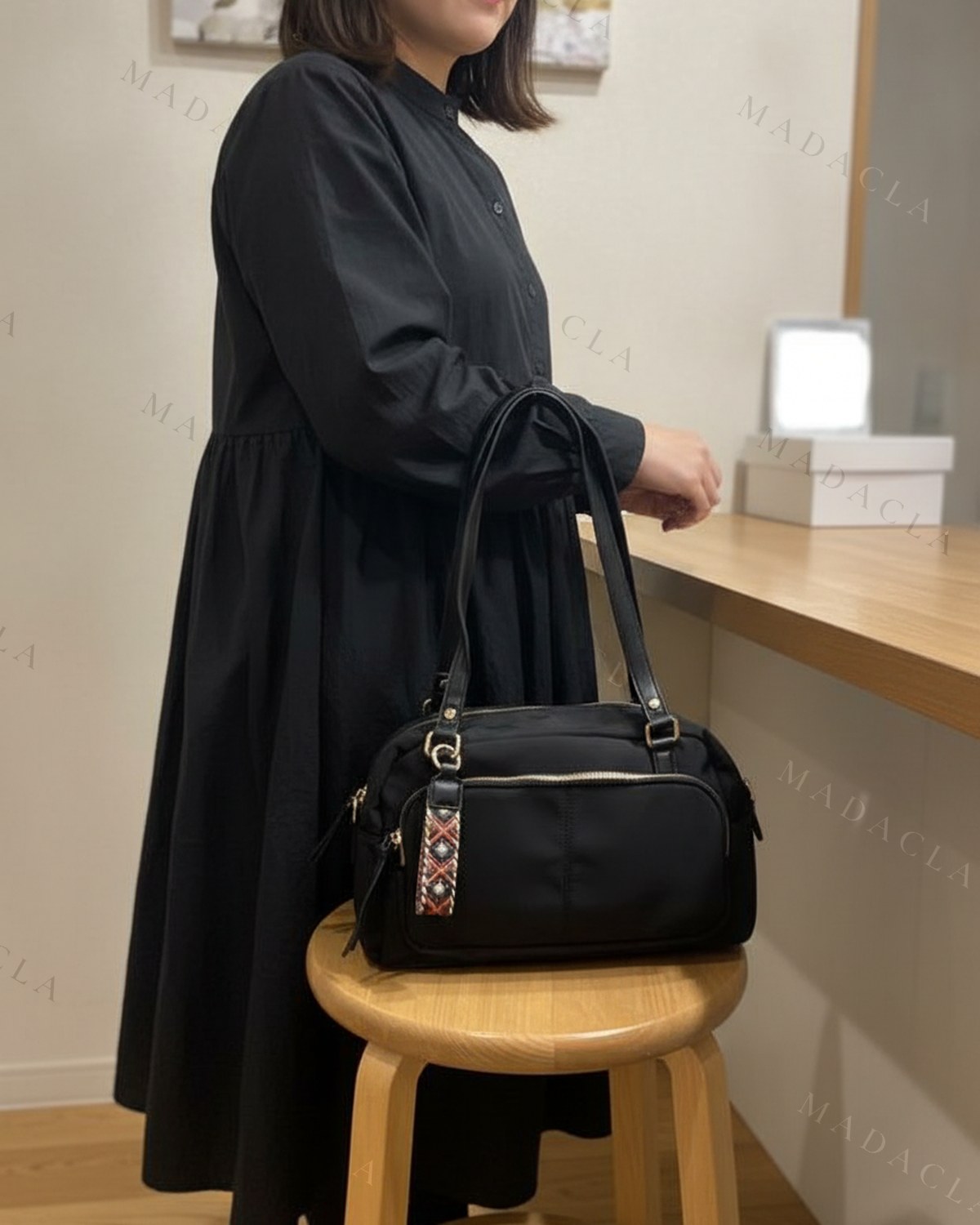 Bolso de nailon negro con forma de bolo, asas de piel, cremallera dorada y bolsillos laterales. Lleva bandolera ancha de estampado geométrico en rojo, blanco y negro.